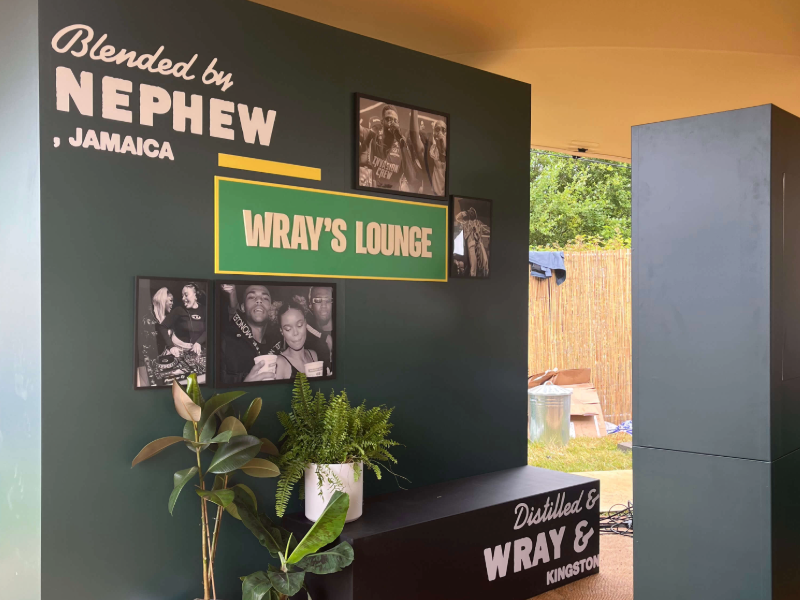 Green Wray's Lounge wall with framed black and white photographs, plants and Blended by Nephew, Jamaica signage