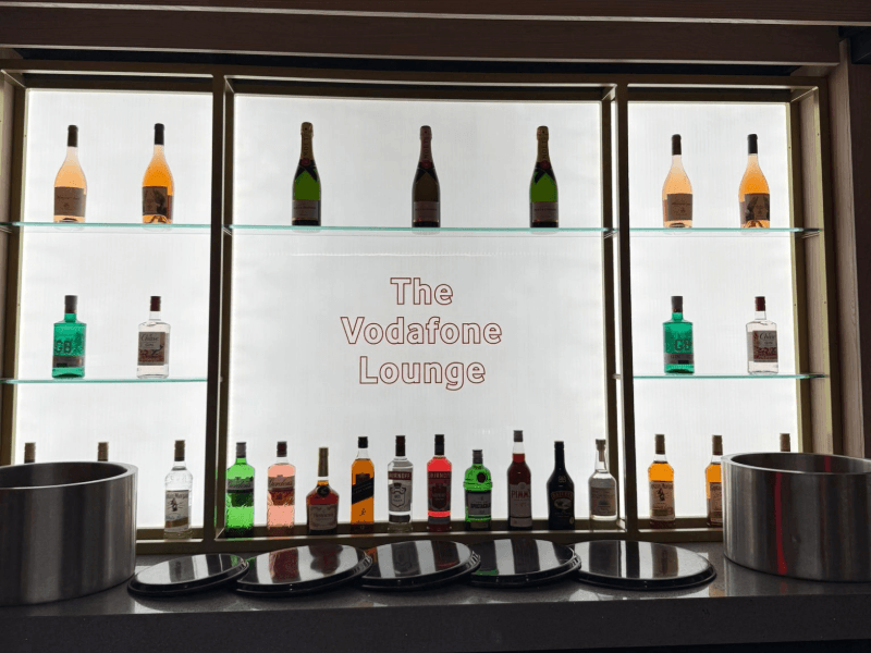 Backlit bar in The Vodafone Lounge with glass shelving, bottles and The Vodafone Lounge signage